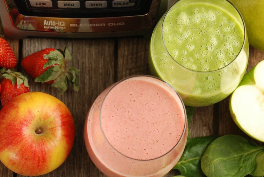 two smoothies, one green and one pink, in glass cups. Other ingredients like apples, strawberries and spinach are next to the cups.