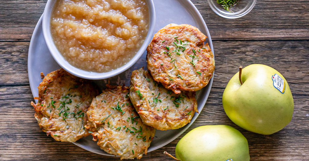 Potato pancakes on a plate with a bowl of homemade applesauce, golden delicious apples are placed next to the plate