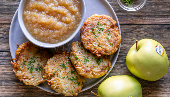 Potato pancakes on a plate with a bowl of homemade applesauce, golden delicious apples are placed next to the plate
