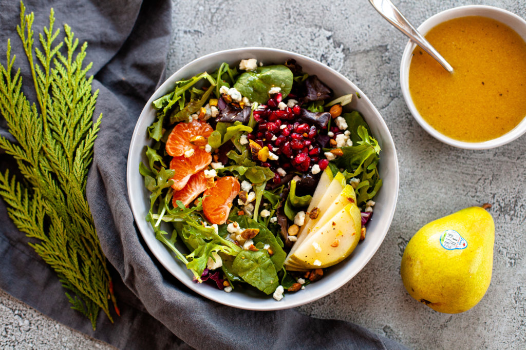 A bowl of winter pear salad with a whole pear and greenery on the table beside the bowl.