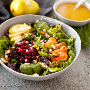 winter pear salad in bowl with dressing in background. On top of the lettuce are pomegranate seeds, sliced pears and peeled mandarine oranges