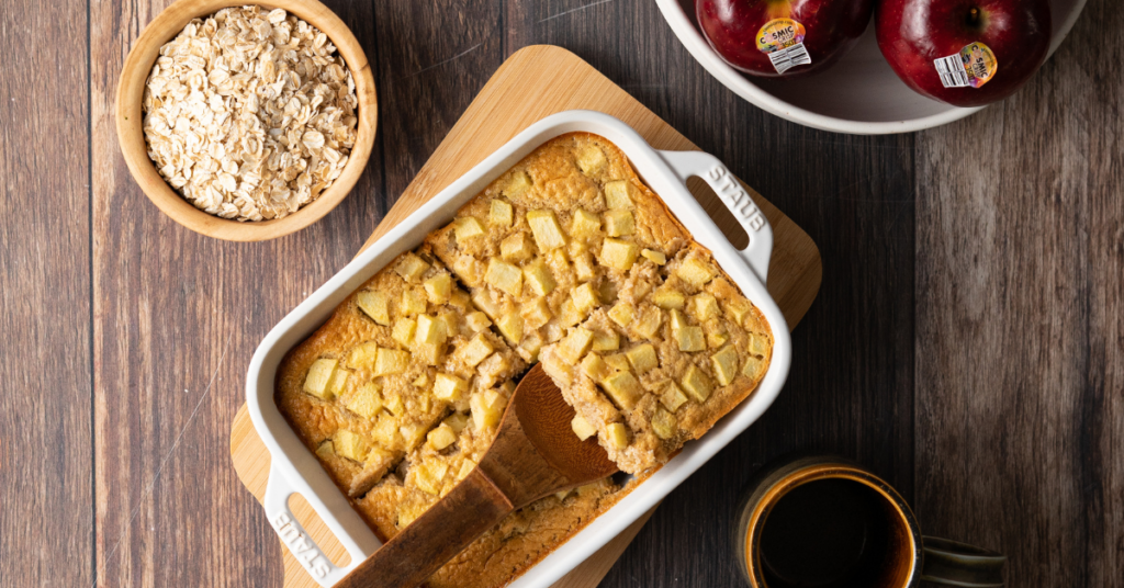 Baked oats with Cosmic Crisp apples in a white casserole dish next to raw oats and a bowl of apples.