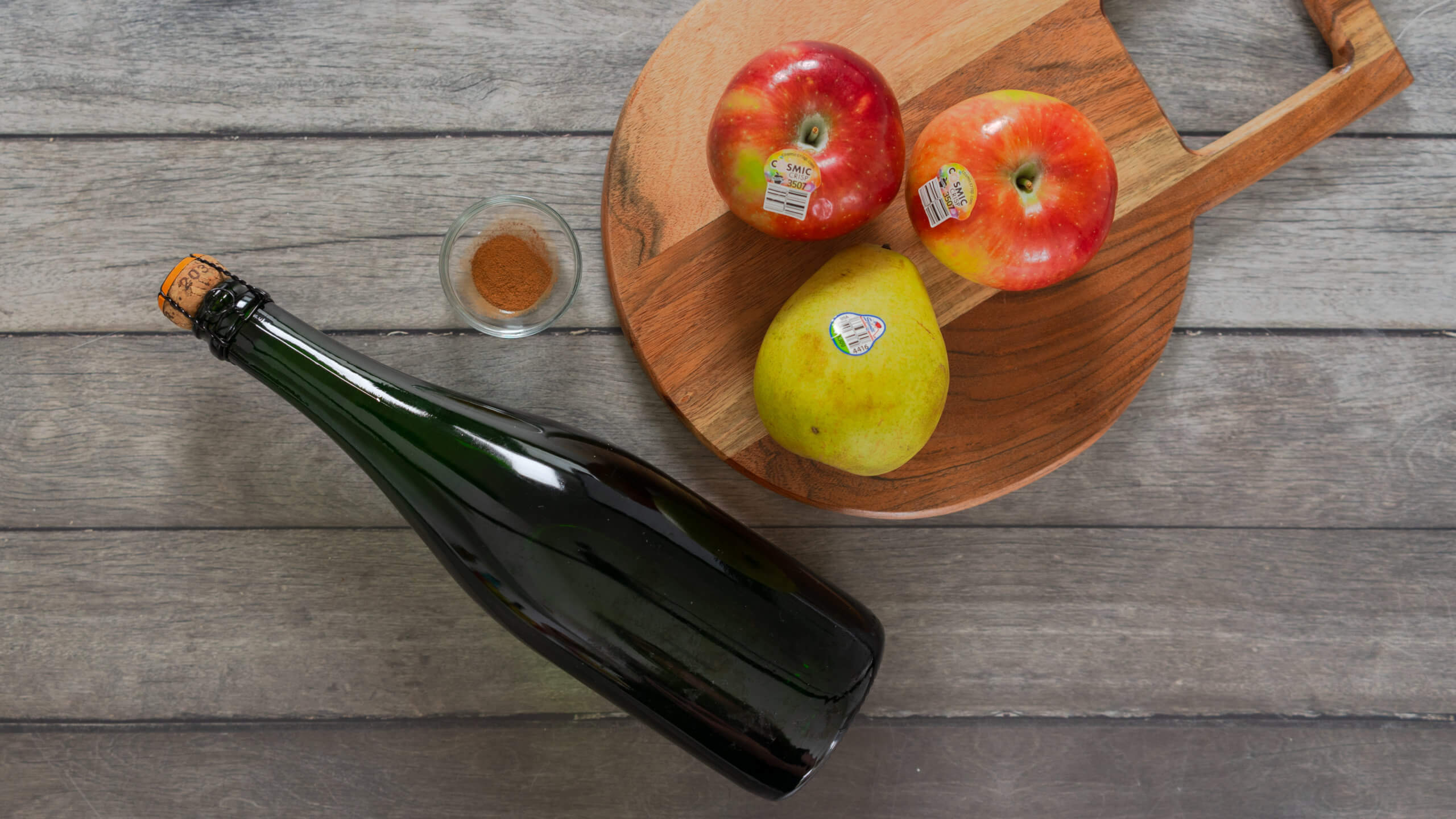 A bottle of champagne next to a cutting board with whole apples