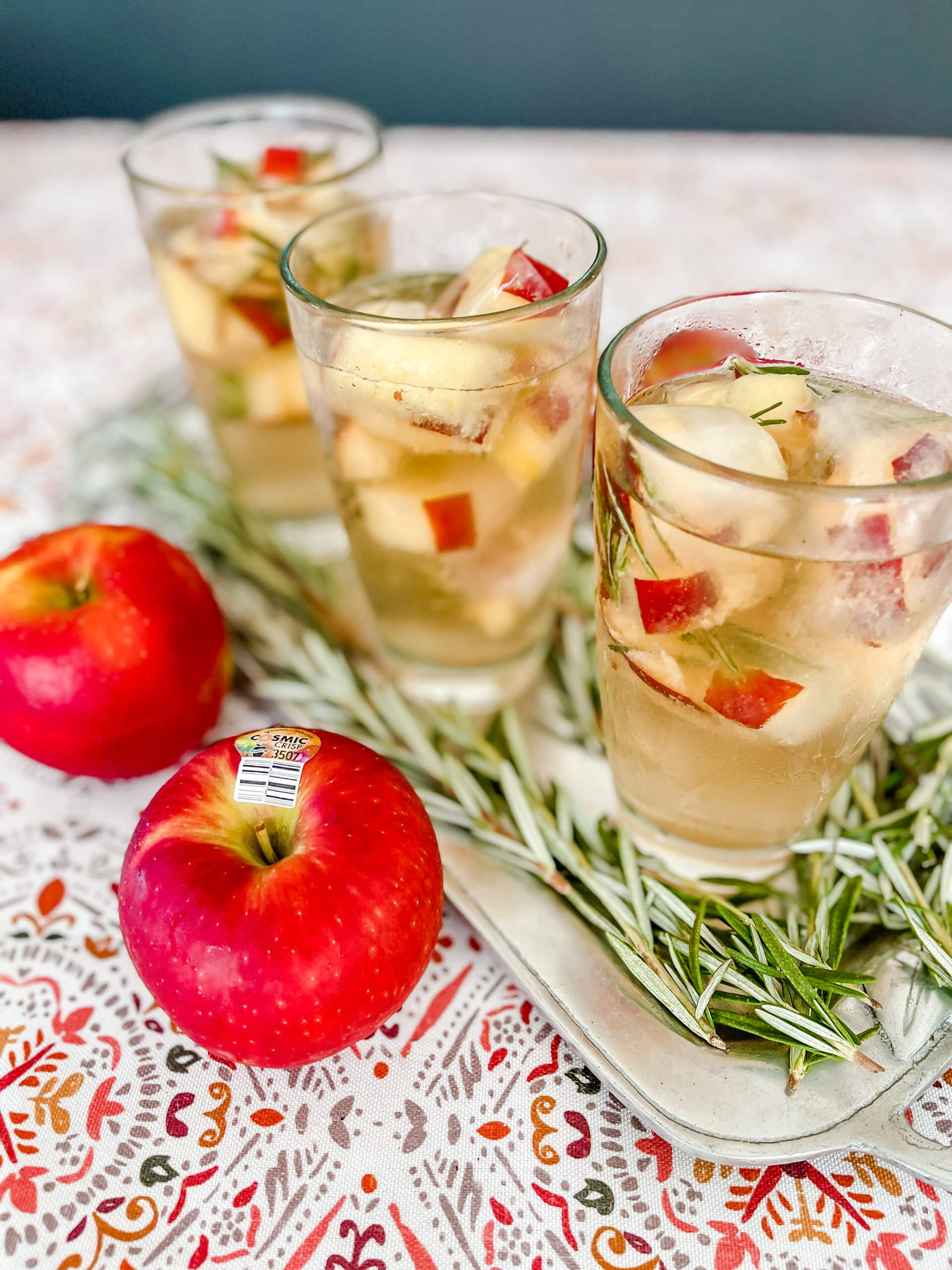 Glasses filled with Ginger Apple Smash ice cubes with Cosmic Crisp apples and sprigs of rosemary on the side.