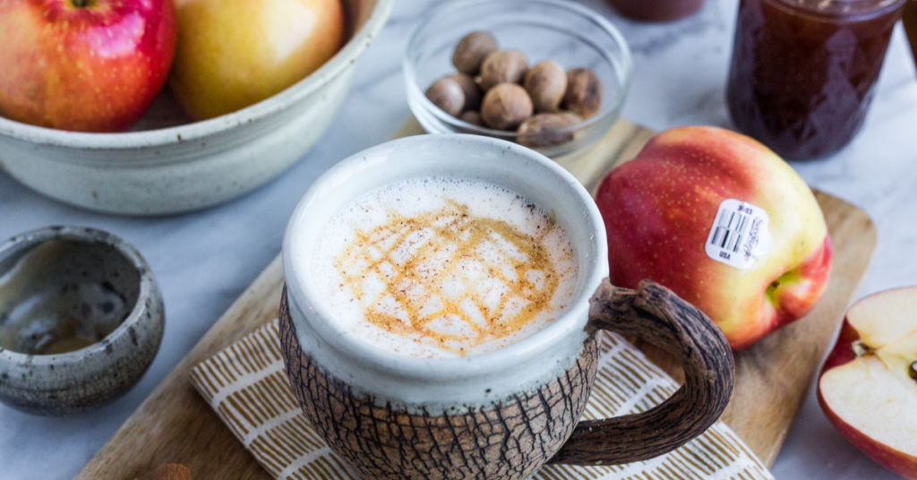 An Apple Crisp Macchiato displayed with whole apples and nutmeg spices