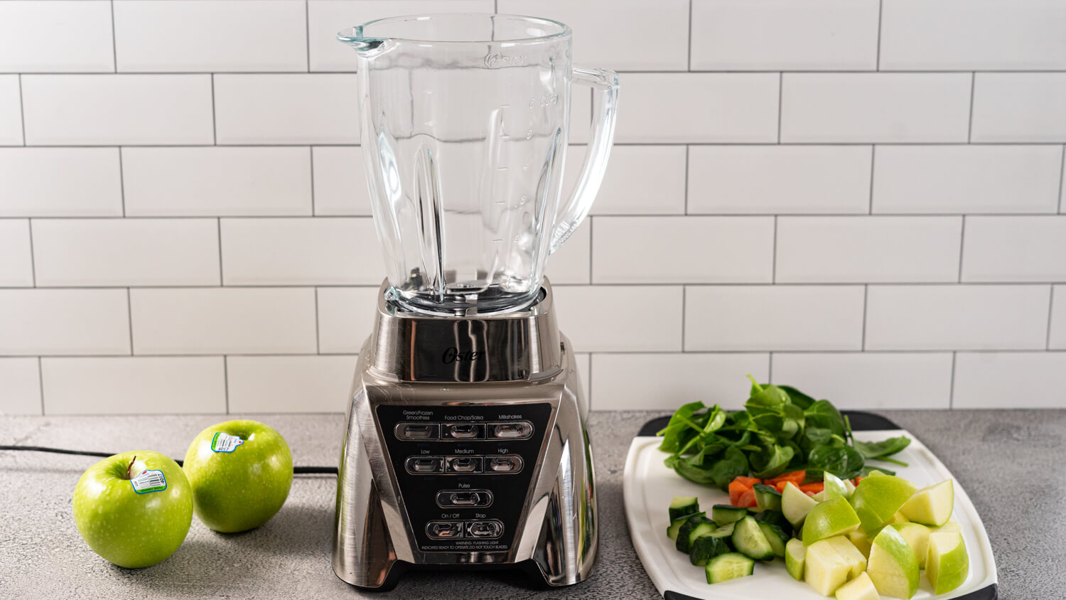 A blender with ingredients beside it to make a Green Apple Smoothie (spinach, apples, carrots, cucumbers).