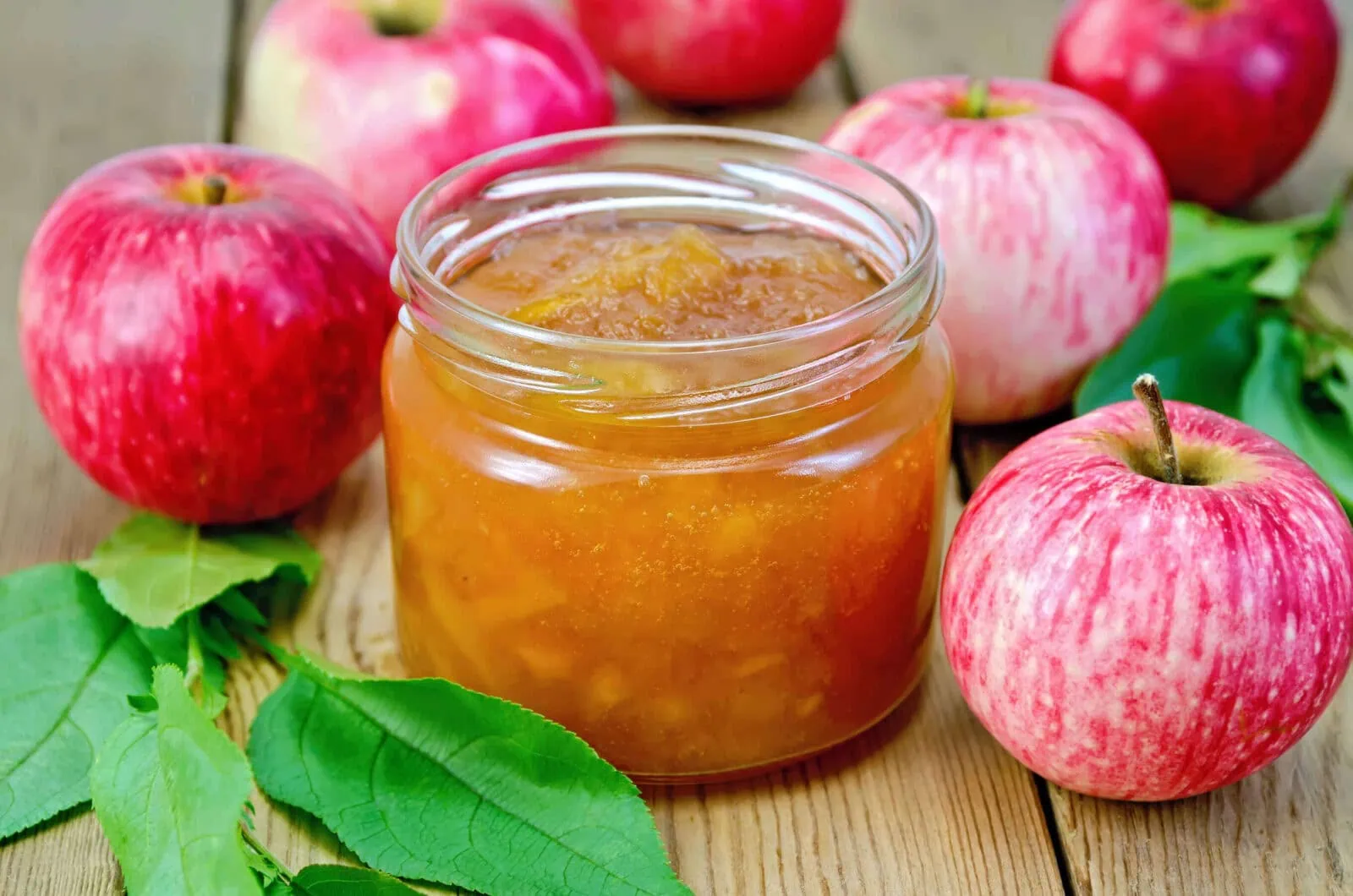 Homemade caramel apple jam in a glass jar with fresh apples and green leaves.