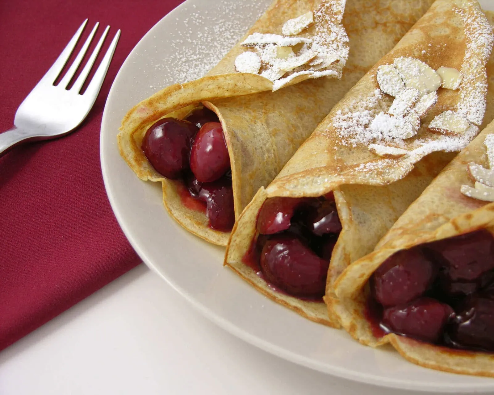 Three cherry-filled crepes dusted with powdered sugar on a white plate.