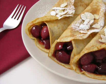 Three cherry-filled crepes dusted with powdered sugar on a white plate.