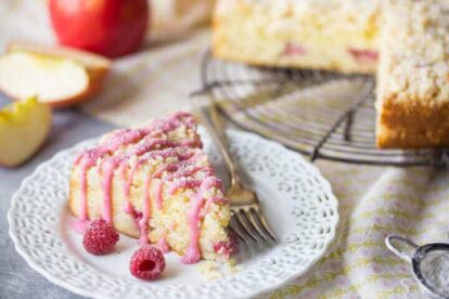 Slice of apple raspberry crumb cake with pink glaze and fresh raspberries on a white plate.