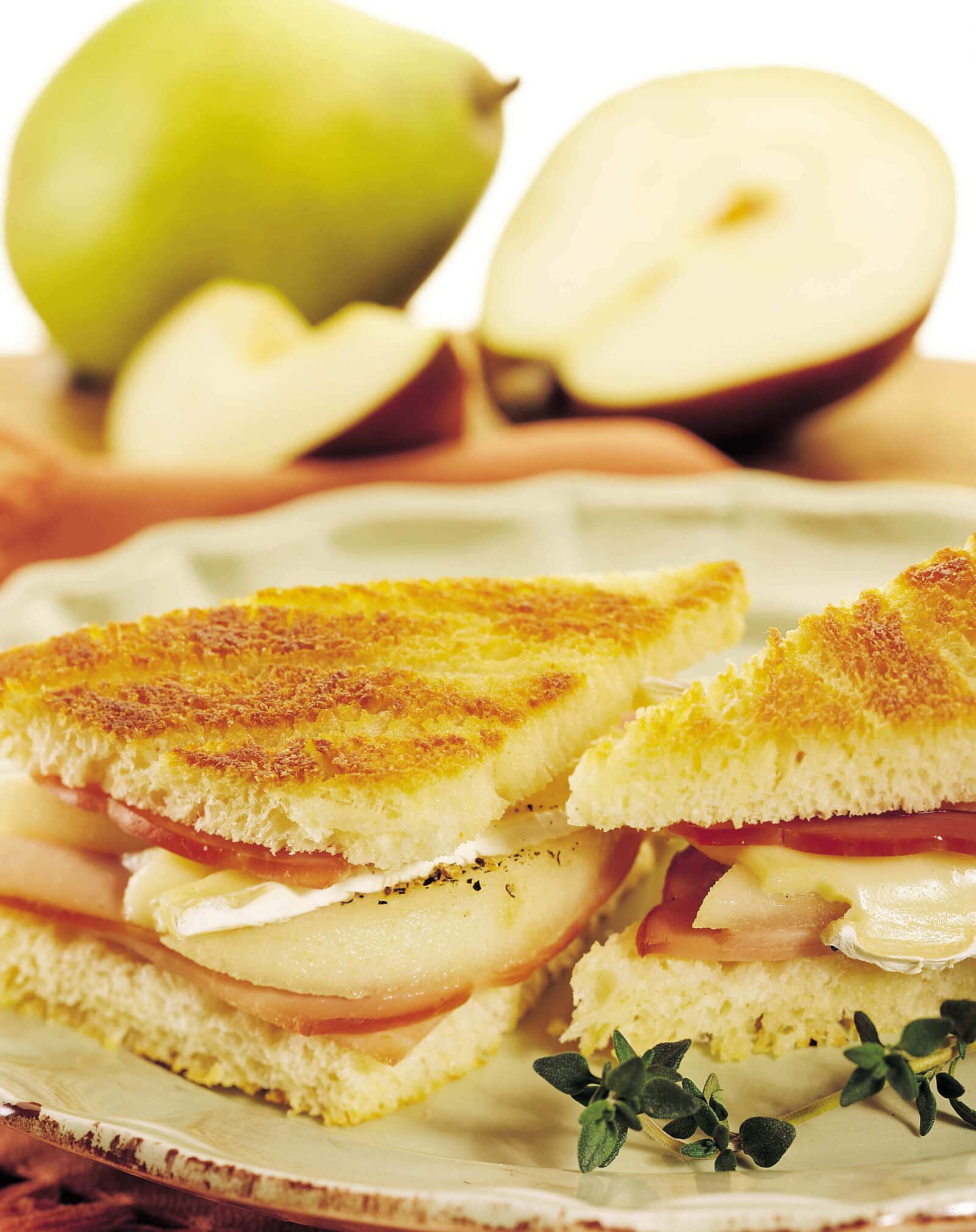 Close-up of Brie, ham, and pear panini sandwiches on a plate with fresh pears in background.