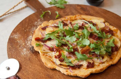 Apple, pear, prosciutto pizza topped with arugula on a wooden serving board.