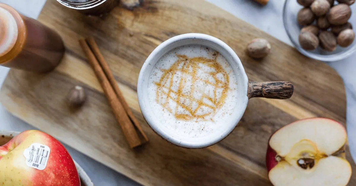 Apple crisp macchiato with caramel drizzle in a mug on a wooden tray with apples and cinnamon sticks.