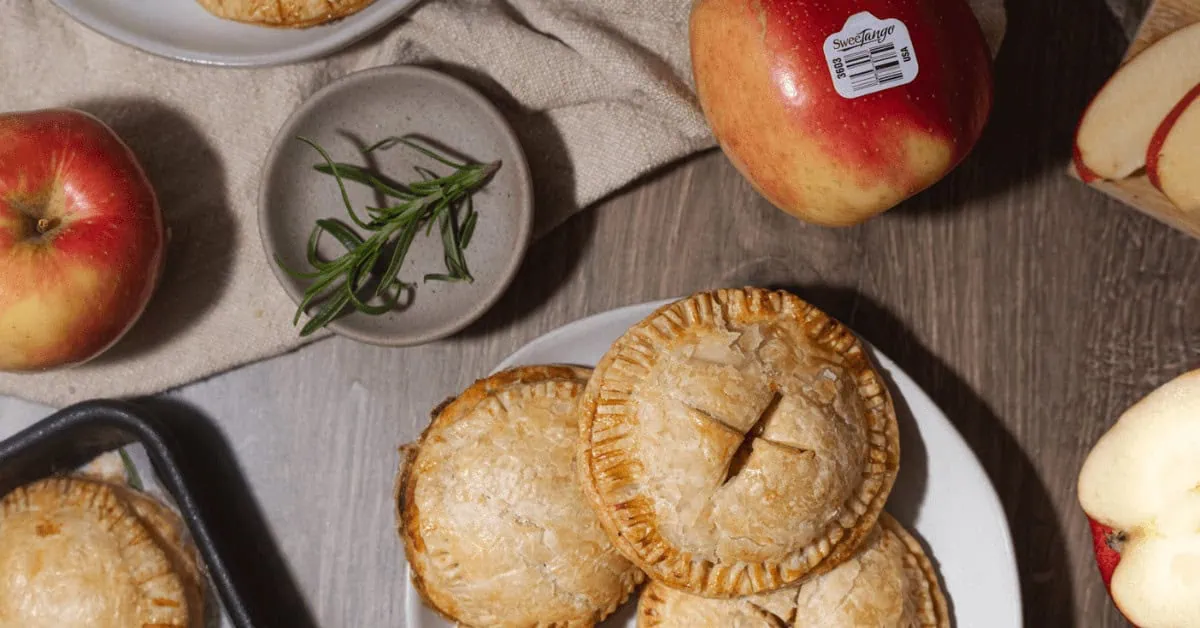 Mini apple and sweet potato pies on a white plate with apples and herbs nearby.
