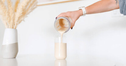 Pouring a creamy smoothie into a glass on a white countertop with decorative pampas grass.