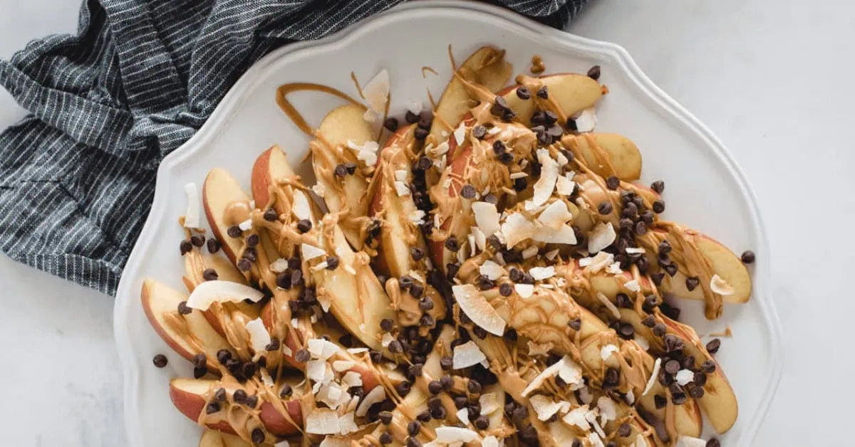 Apple nachos topped with caramel, chocolate chips, and coconut flakes on a white plate.