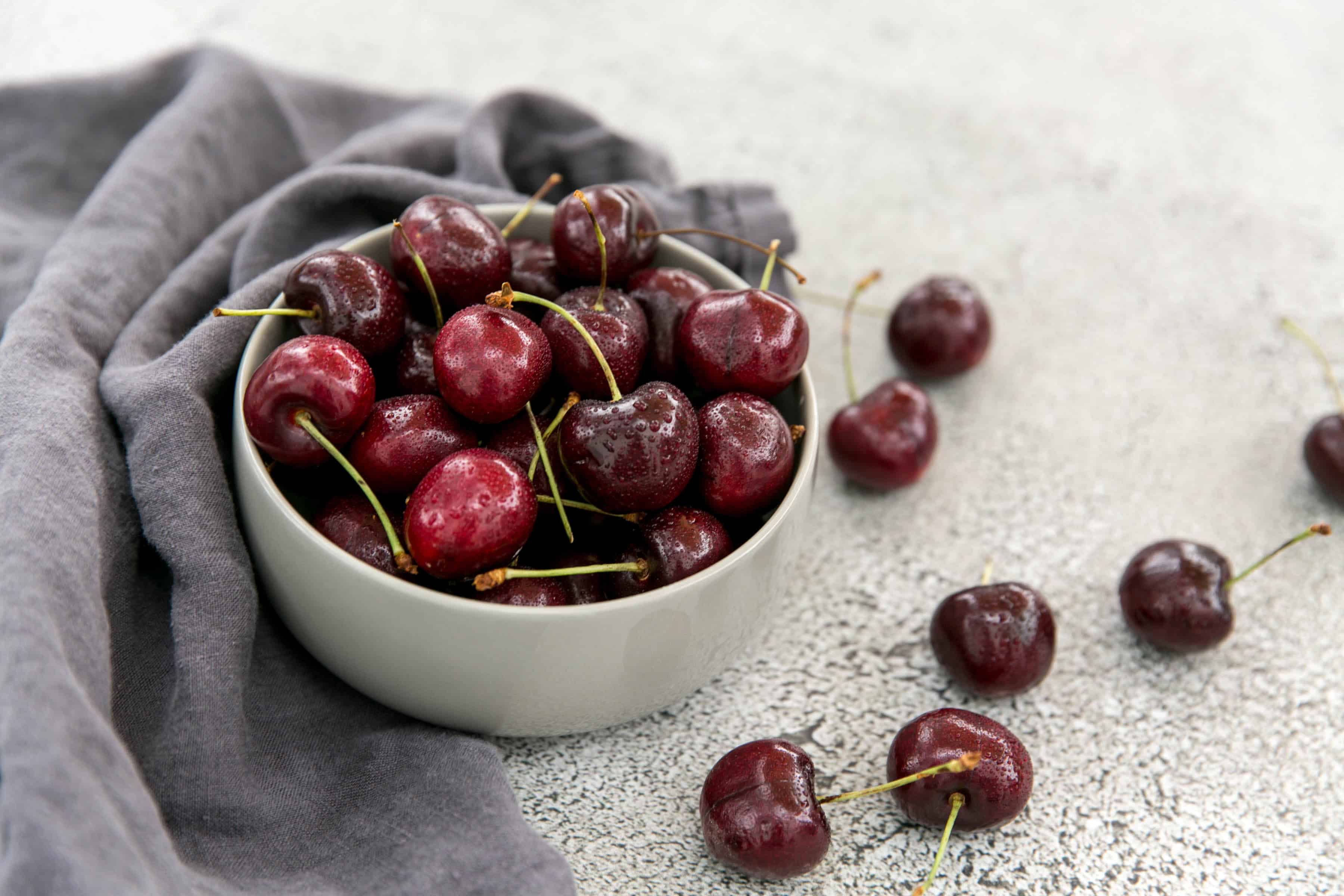 Fresh, ripe cherries in a white bowl on a textured surface with a gray cloth.