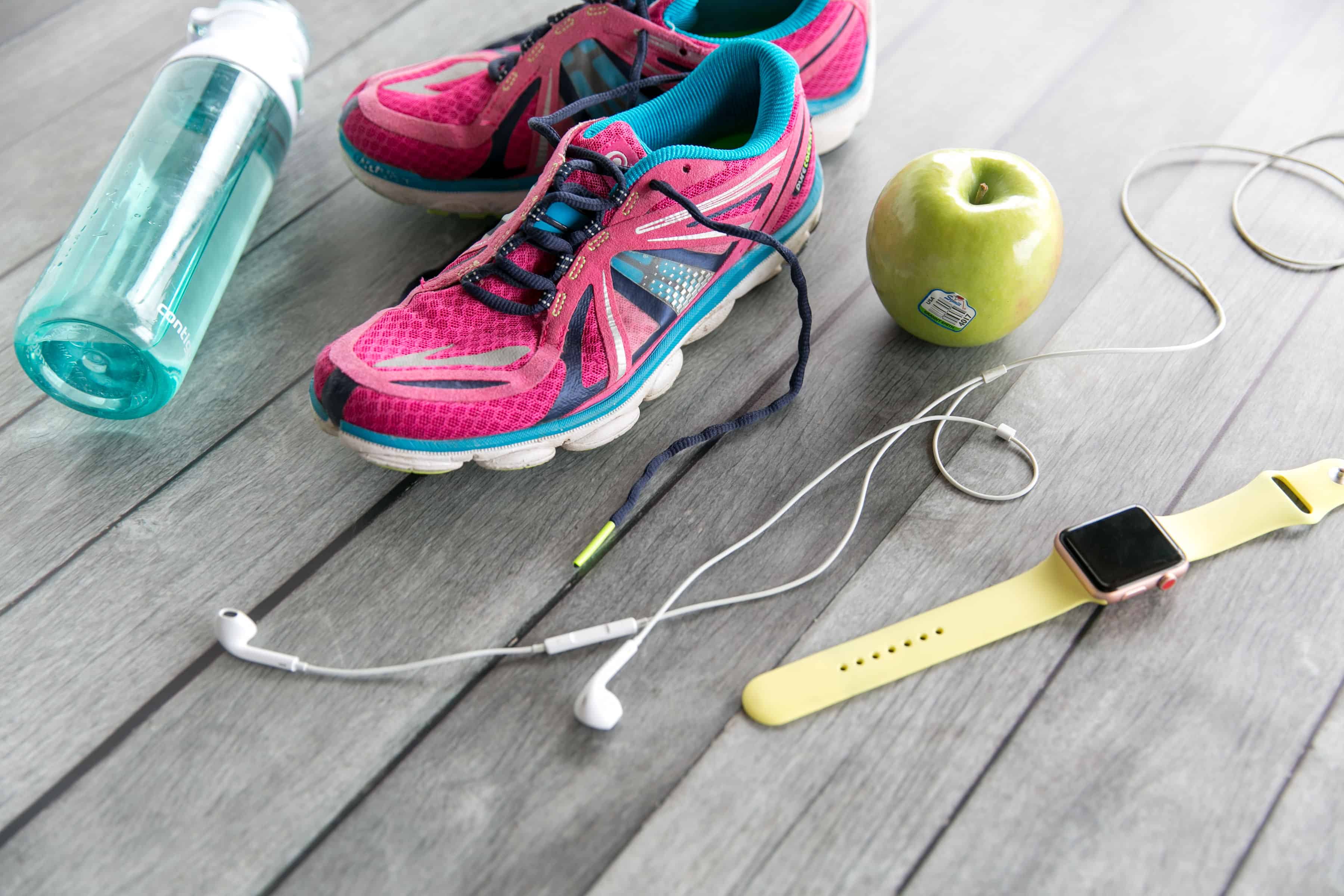Running shoes, apple, water bottle, smartwatch, and earphones on wooden floor.