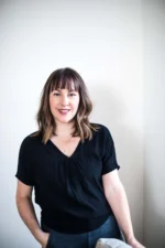 Smiling woman in black top standing against a plain wall.