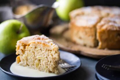 Slice of Irish apple cake with whiskey hard sauce on a black plate with green apples in background.