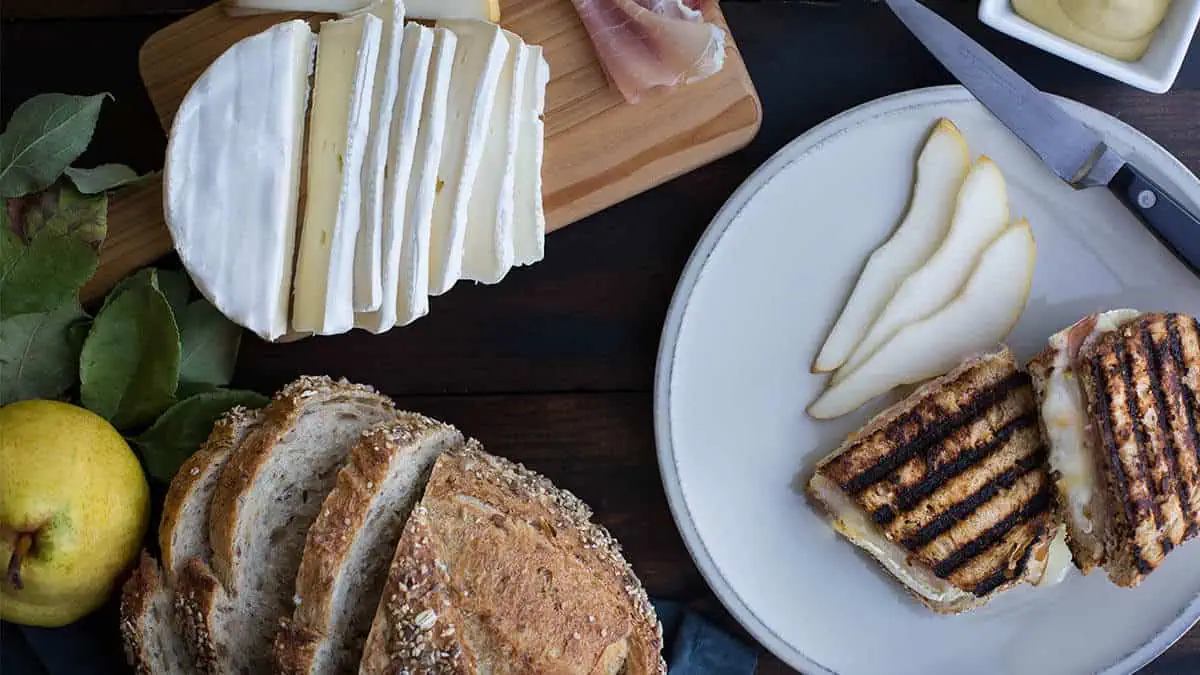 Grilled pear, prosciutto, and brie panini with fresh pears and cheese on a white plate.