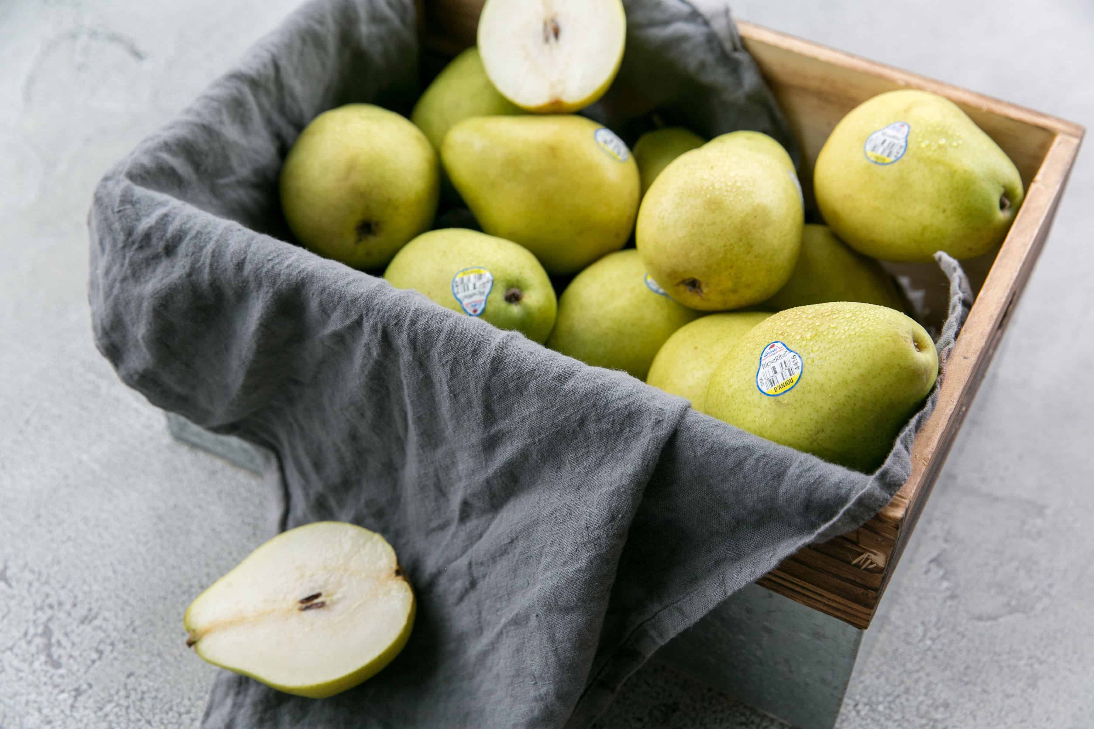 Fresh green pears in a wooden basket with one cut open, on a gray surface.