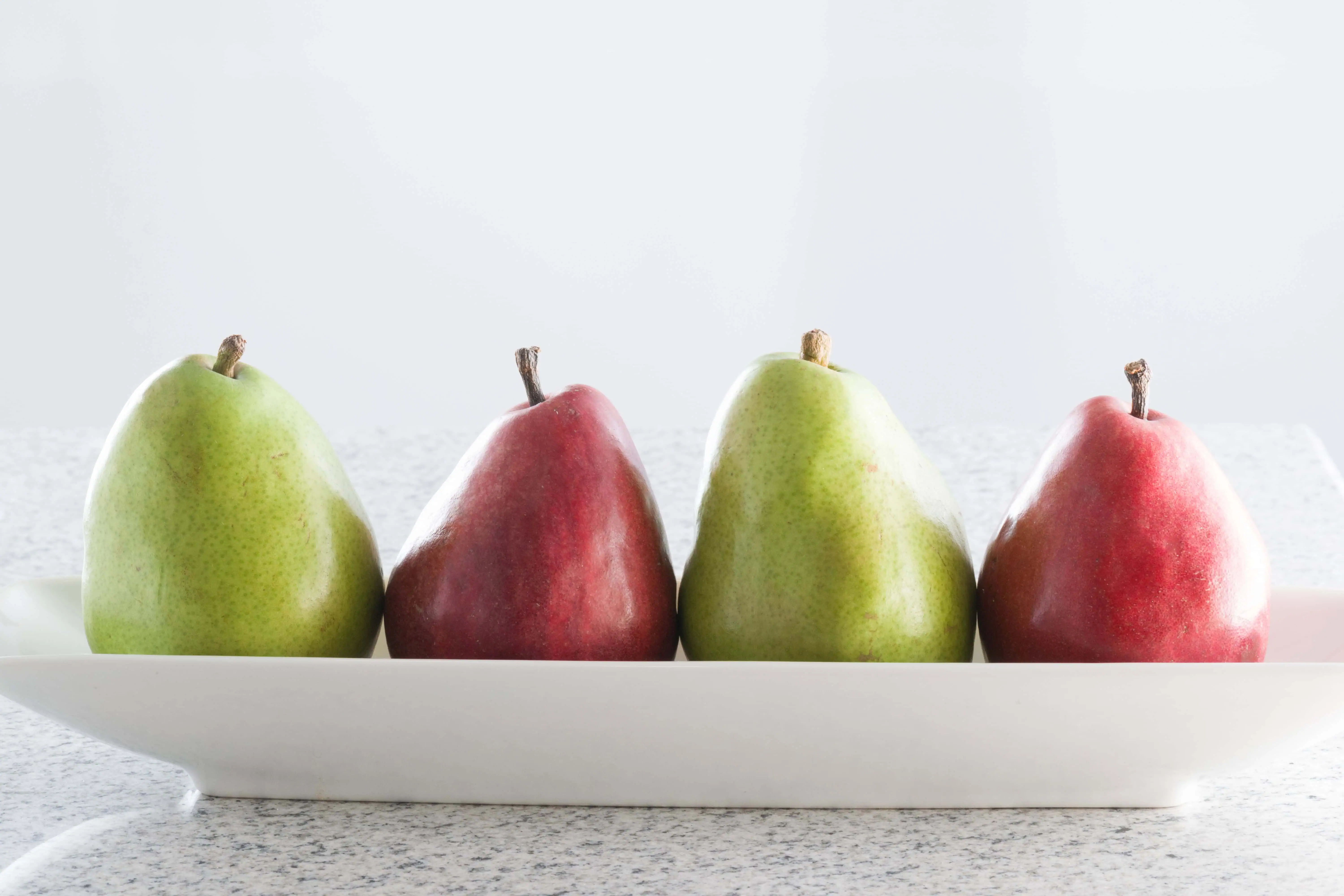 Fresh green and red pears on a white platter for ripening or snacking.