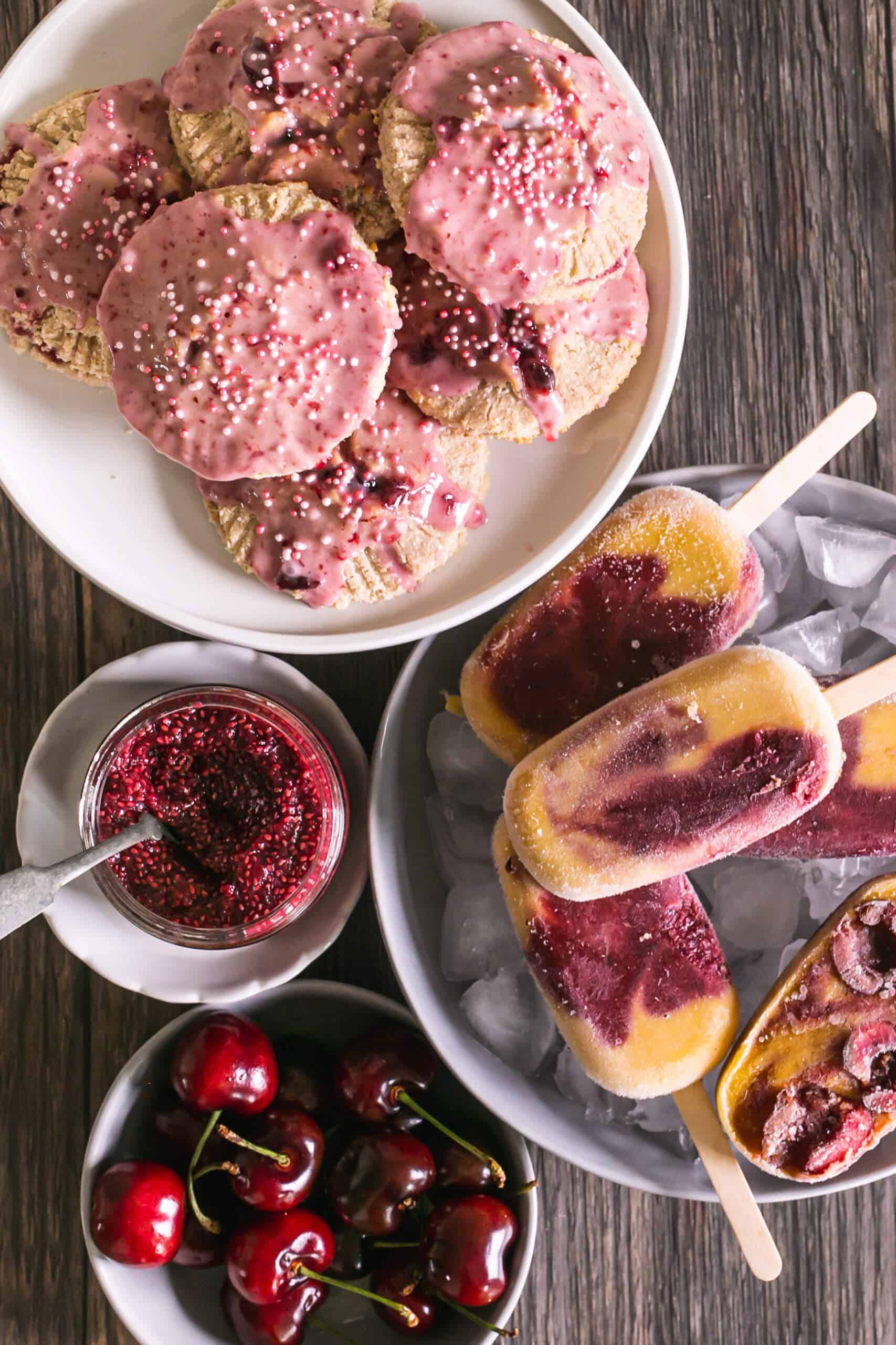 Assorted cherry-flavored popsicles, cherry bowl, and cherry cookies on a wooden table.