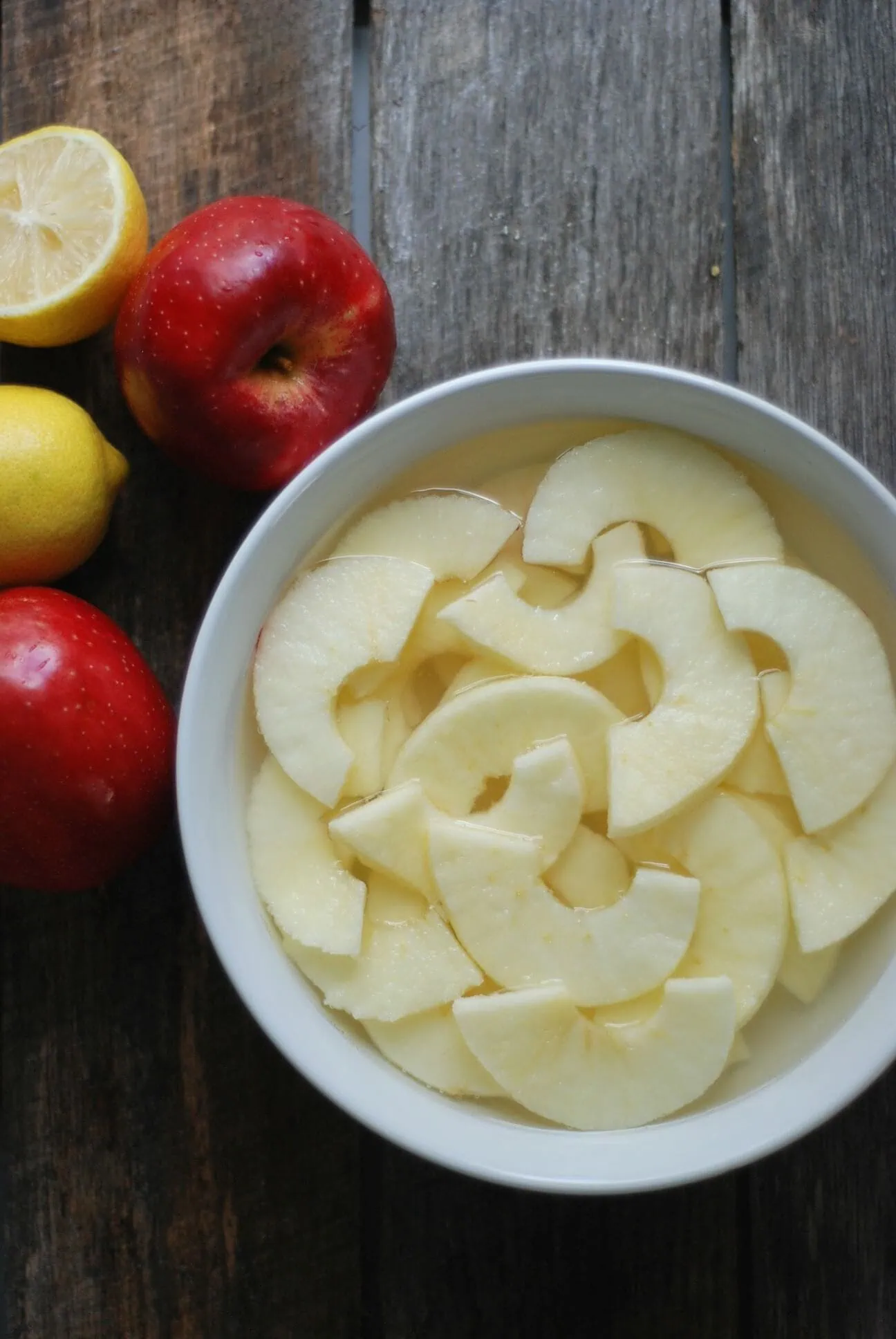 Sliced apples in a white bowl with whole apples and lemons on a wooden surface.