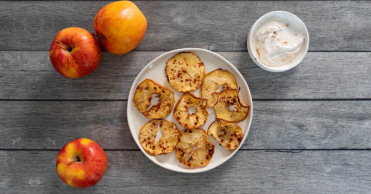 Fresh apple chips on a white plate with whole apples on a wooden surface.