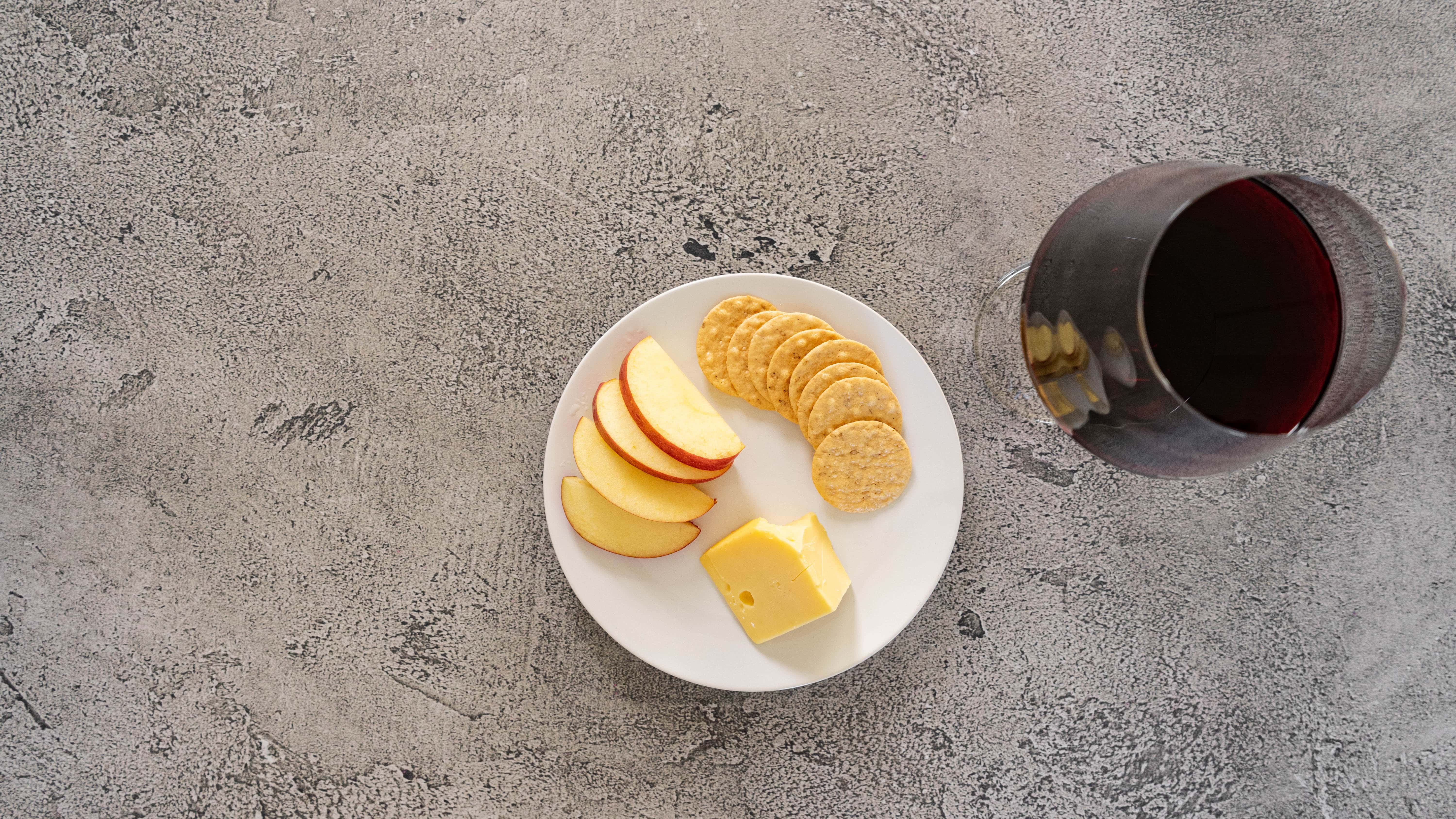 Apple slices, cheese, crackers, and a glass of red wine on a gray surface.