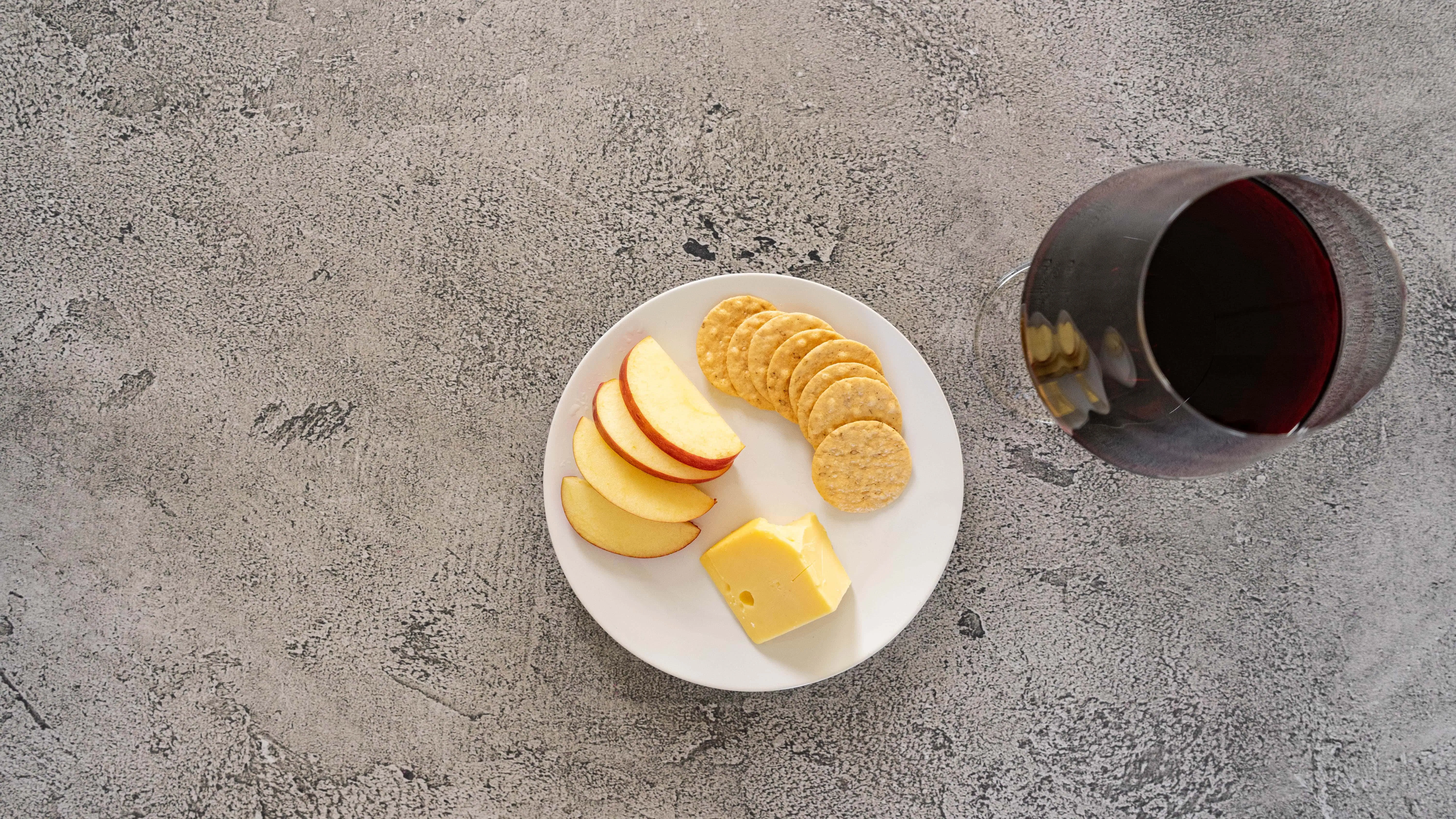 Apple slices, cheese, crackers, and a glass of red wine on a gray surface.