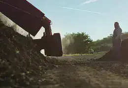 Farmer inspecting soil on sustainable farm with machinery in background.