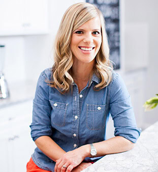 Smiling woman in denim shirt sitting at kitchen counter with plants.