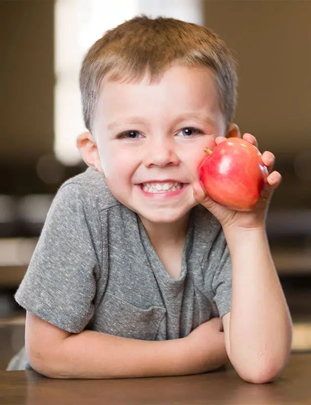 Young boy smiling and holding a red apple near his face.