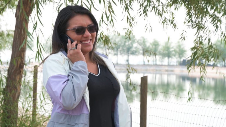 Woman smiling and talking on phone outdoors near water and trees.